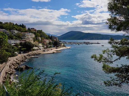 This landscape photograph captures a scenic view looking towards Herceg Novi from the Mirko Komnenović museum gardens, during a bright spring morning in Montenegro. The image prominently features the charming coastal town of Herceg Novi extending along the Adriatic shoreline, with a sweep of colourful buildings and lush greenery along the popular Pet Danica promenade. In the background, the hills of Boka Bay rise gently, while the clear blue waters of the bay are dotted with moored boats and a distant marina, highlighting the picturesque appeal of this part of Montenegro. The photo's vibrant lighting and tranquil atmosphere evoke the serene beauty for which Boka Bay and Herceg Novi are renowned.